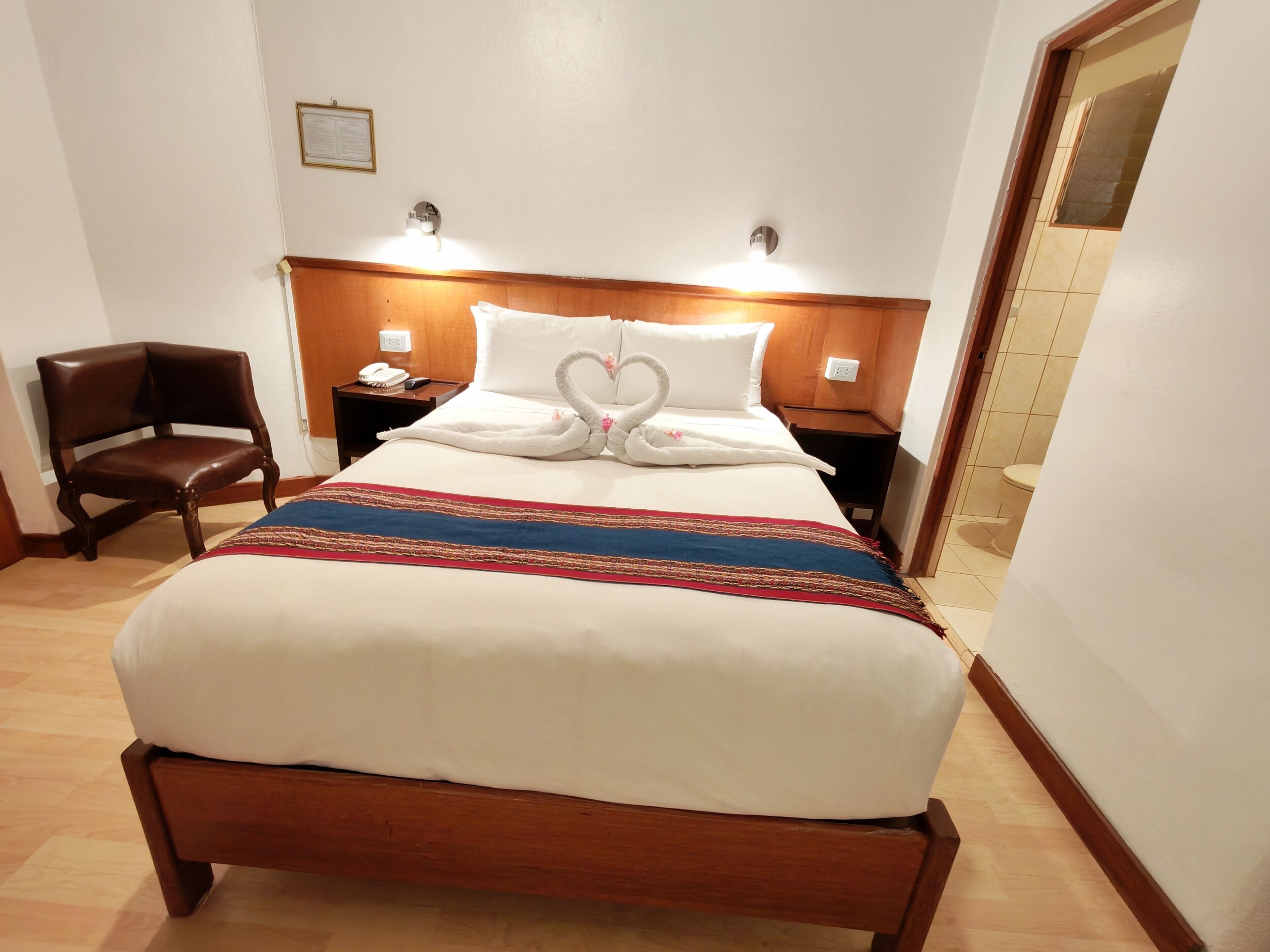 Una habitación de hotel con una cama de madera, una mesita de noche con un teléfono, una silla y una lámpara fijada a la pared.
