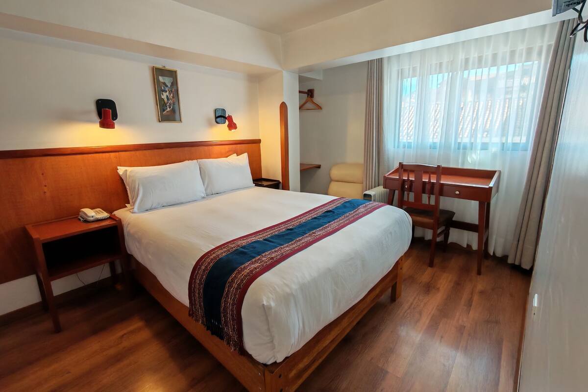 A hotel room with a wooden bed, a desk, a chair, a telephone, and a window with curtains.