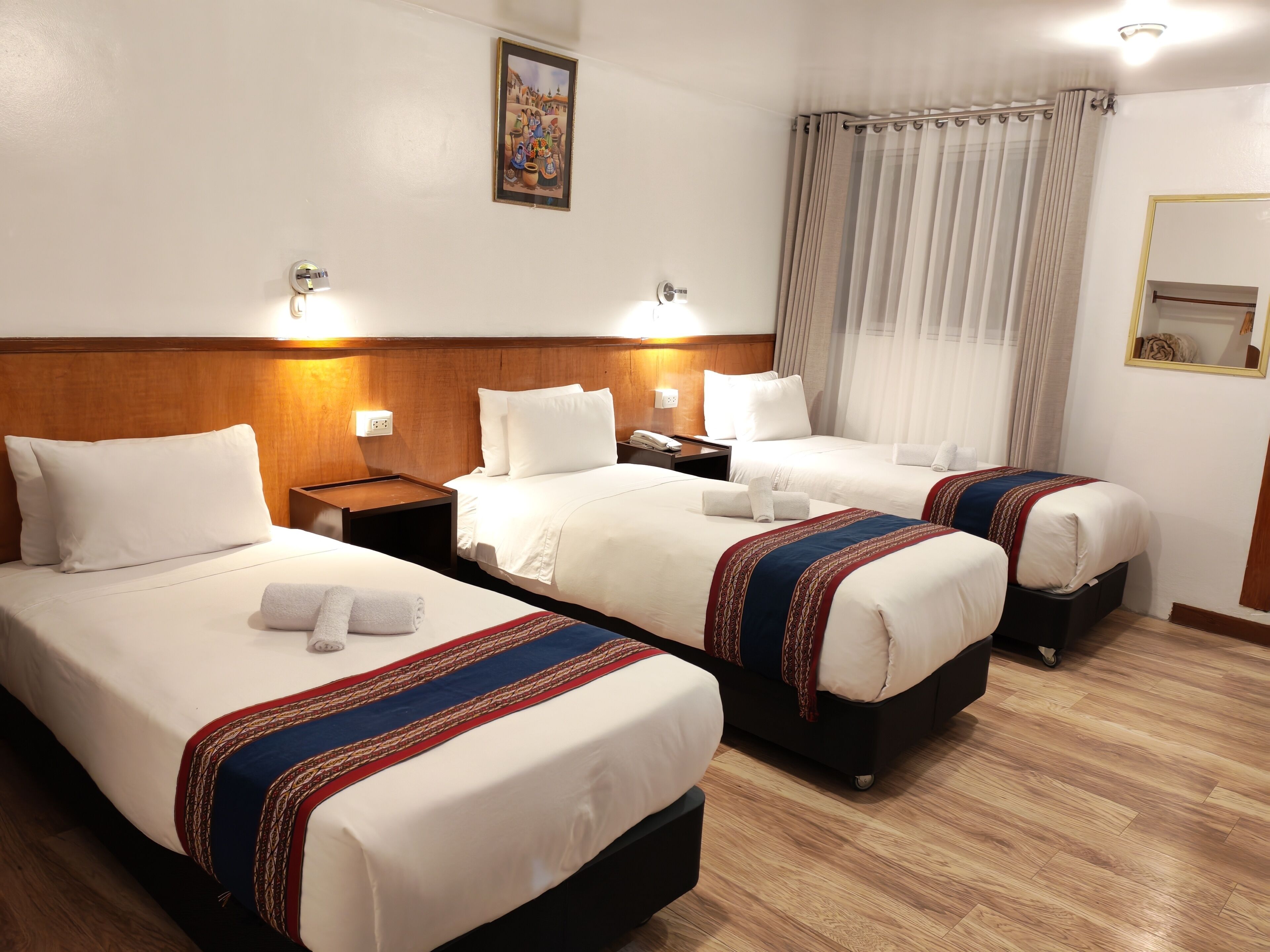A hotel room with three beds, a wooden headboard, a painting on the wall, and a window with curtains.