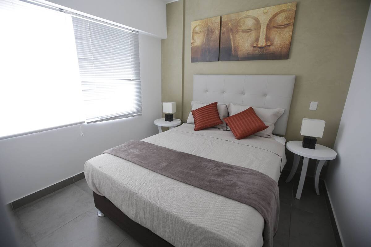 A bedroom with a large bed, two bedside tables, a window with blinds, and a wall with a Buddha painting.