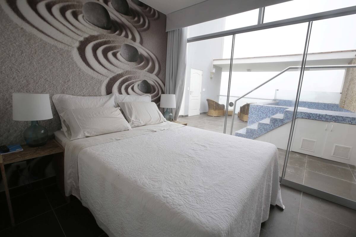 A modern hotel room with a large bed, a nightstand, a lamp, and a view of a pool area through a glass door.