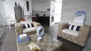Una sala de estar moderna con muebles de mimbre, una mesa de centro con libros y un cuenco decorativo, y una cocina al fondo.