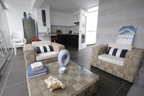 A modern living room with wicker furniture, a coffee table with books and a decorative bowl, and a kitchen in the background.