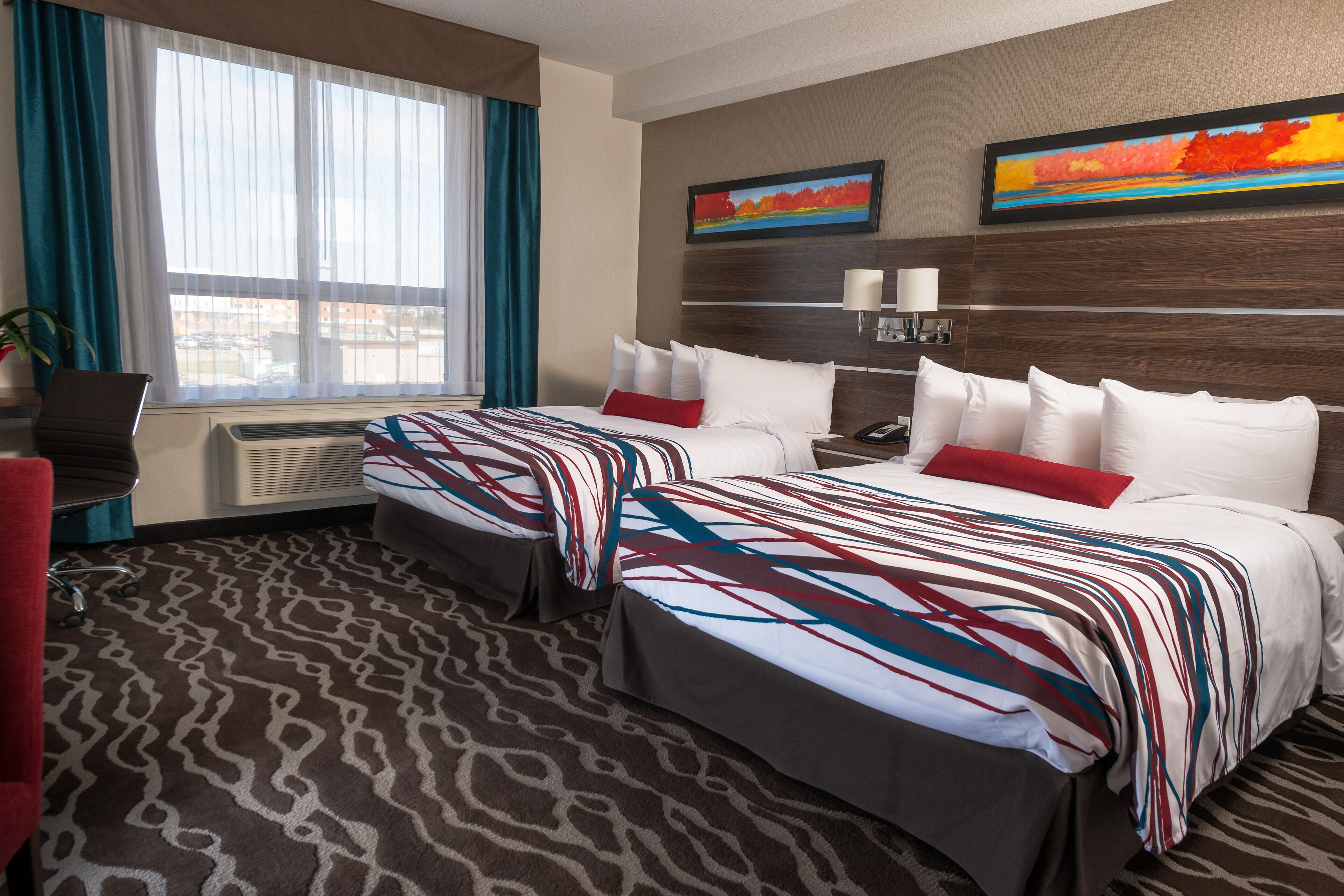 A hotel room with two beds, a desk, and a chair. There are two paintings on the wall and a window with curtains.