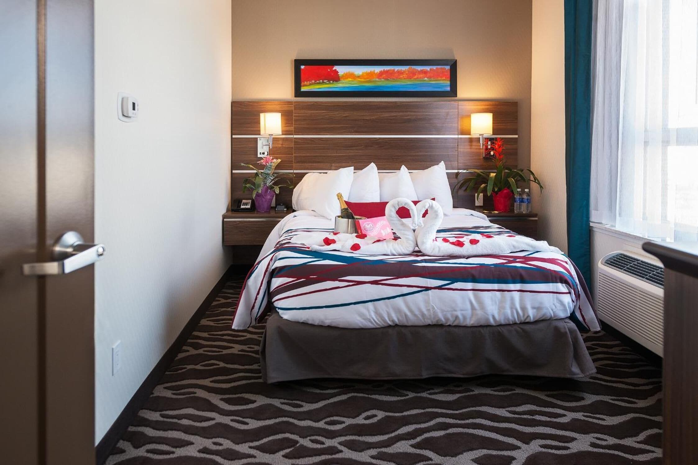A hotel room with a bed, a wall-mounted TV, a bedside table with a lamp, and a window with curtains.