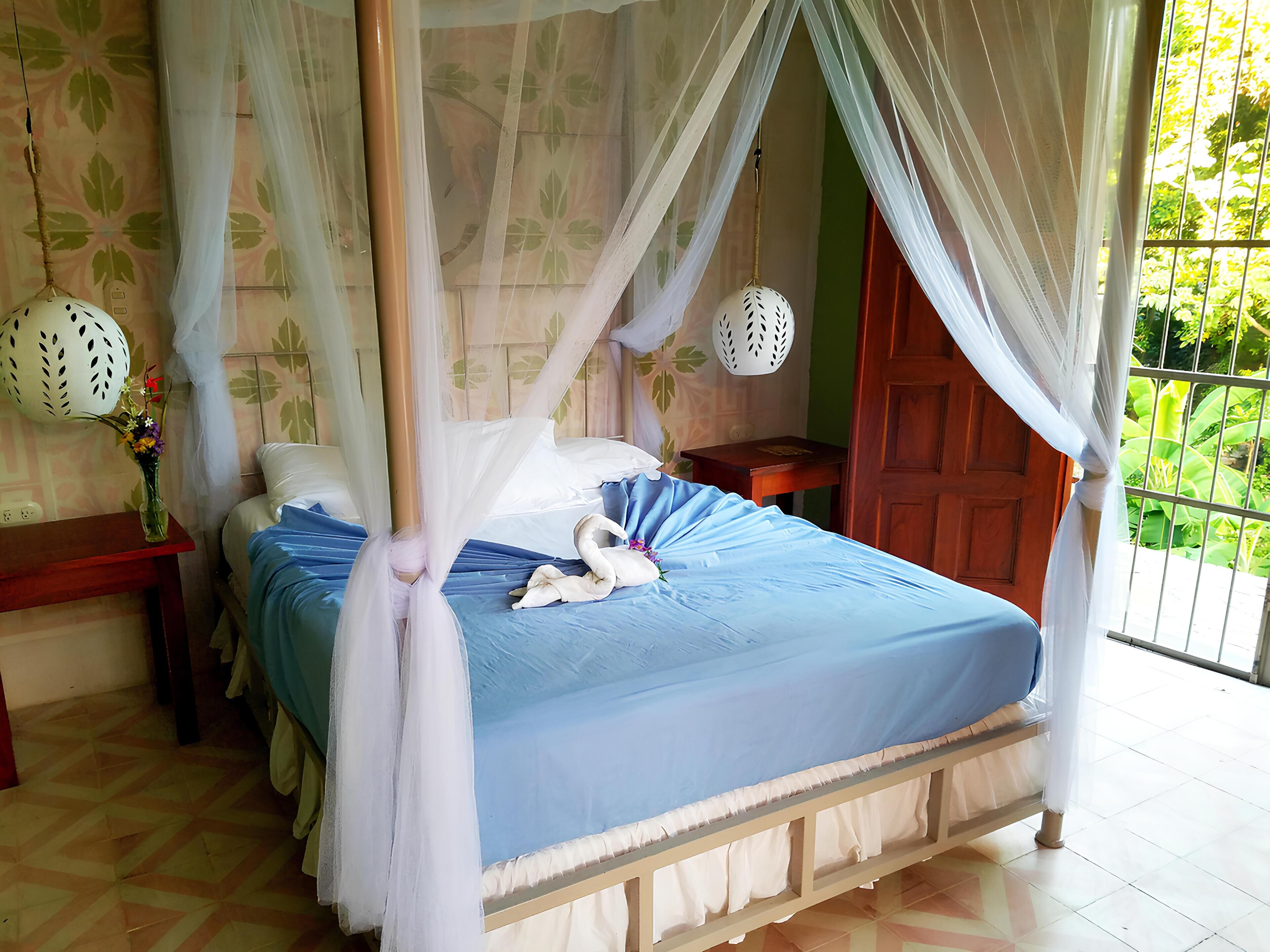 A four-poster bed with a canopy, a bedside table with a vase of flowers, and a view of greenery through a window.