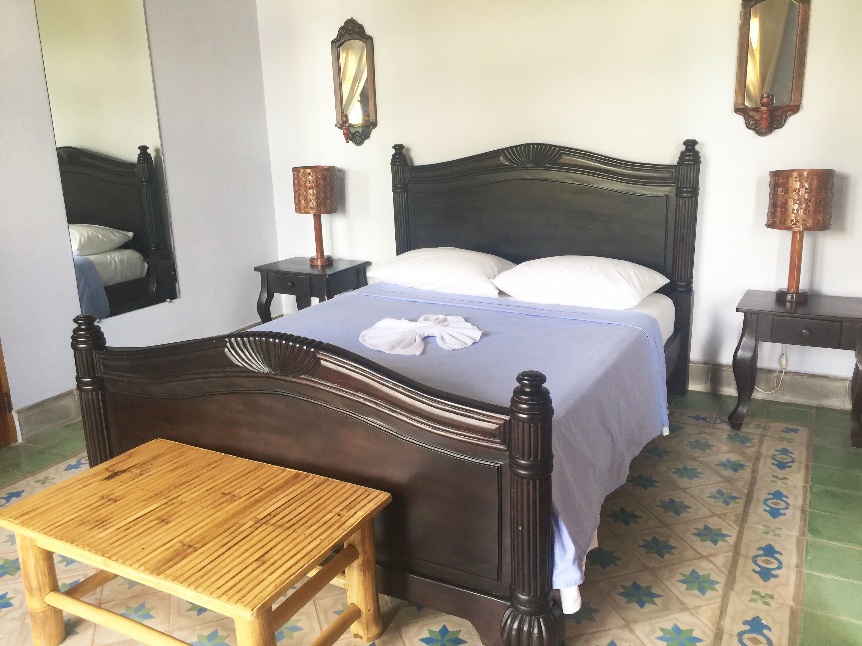 A neatly made bed with a blue bedspread and white pillows, flanked by two bedside tables with lamps, a wooden bench, and a large mirror on the wall.