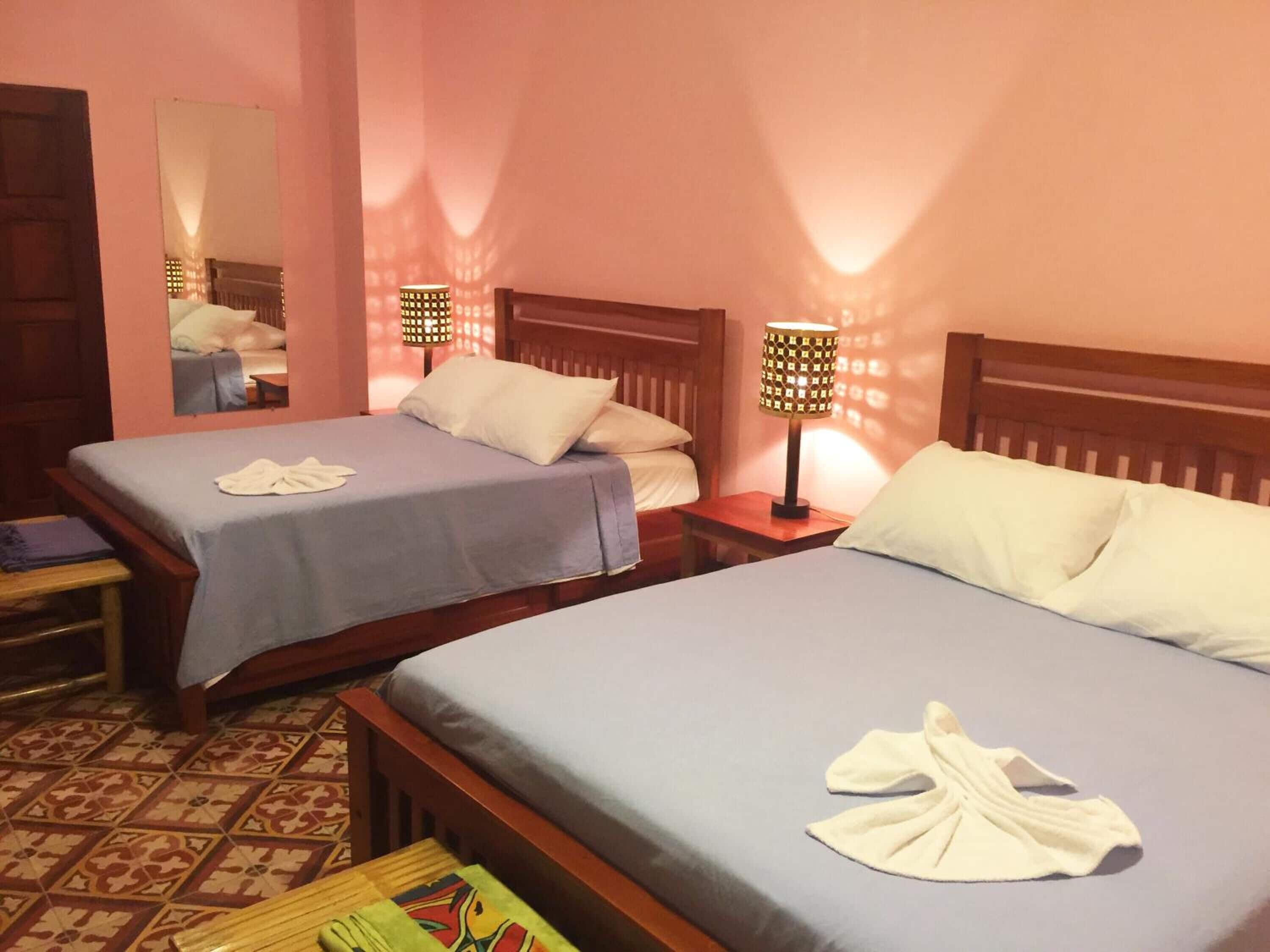 A hotel room with two beds, each with a blue bedspread and white pillows. There are two bedside lamps with patterned shades. A wooden bench is placed between the beds, and a small table with a book is visible.