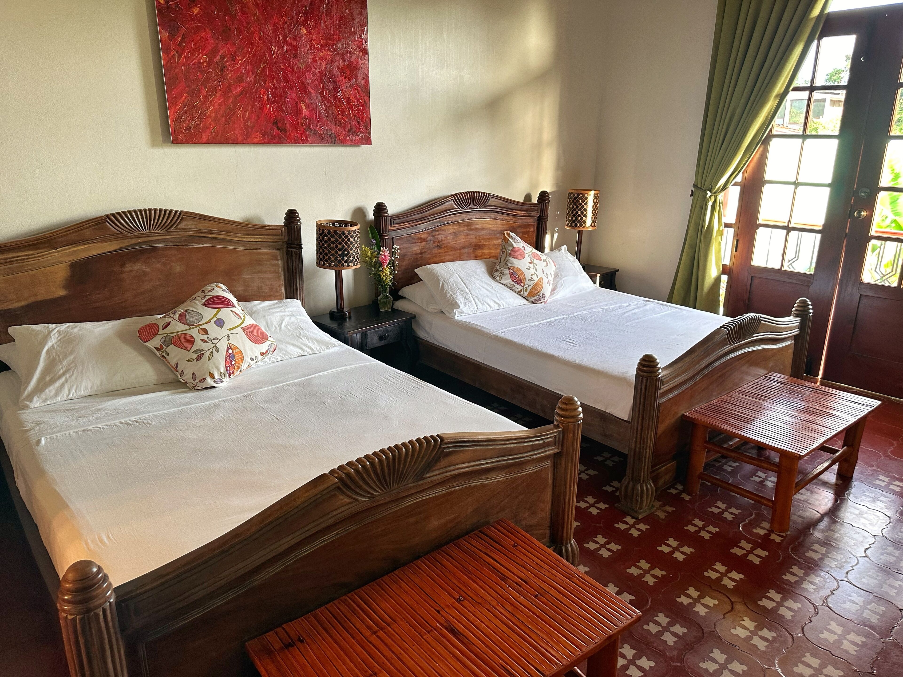 Dos camas de madera con sábanas blancas y almohadas estampadas, una mesita de madera y un suelo con estampado rojo.