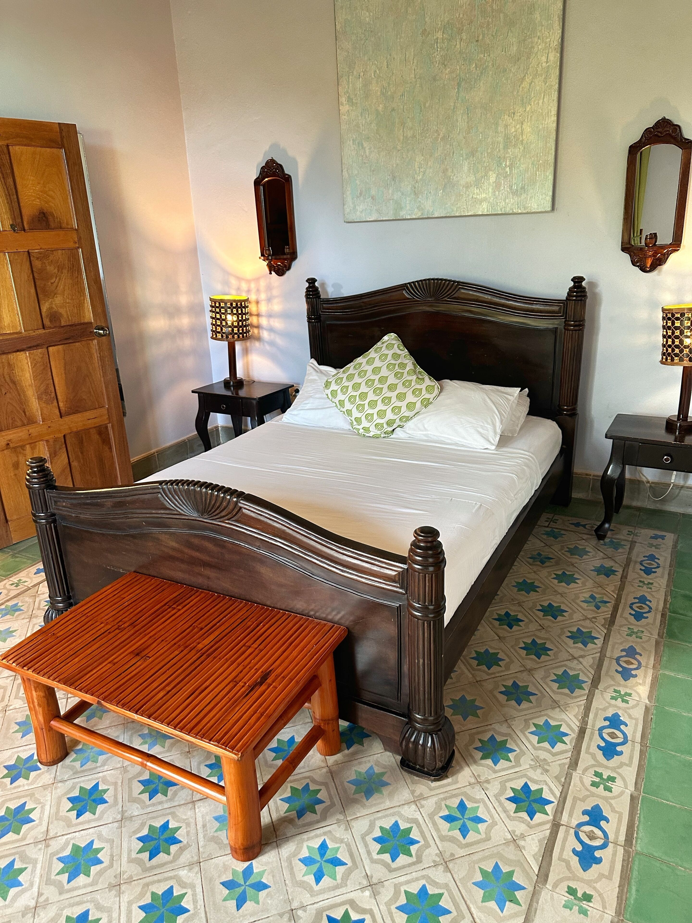 Una cama bien hecha con un cabecero de madera, una mesita de noche con una lámpara, y un banco frente a la cama.