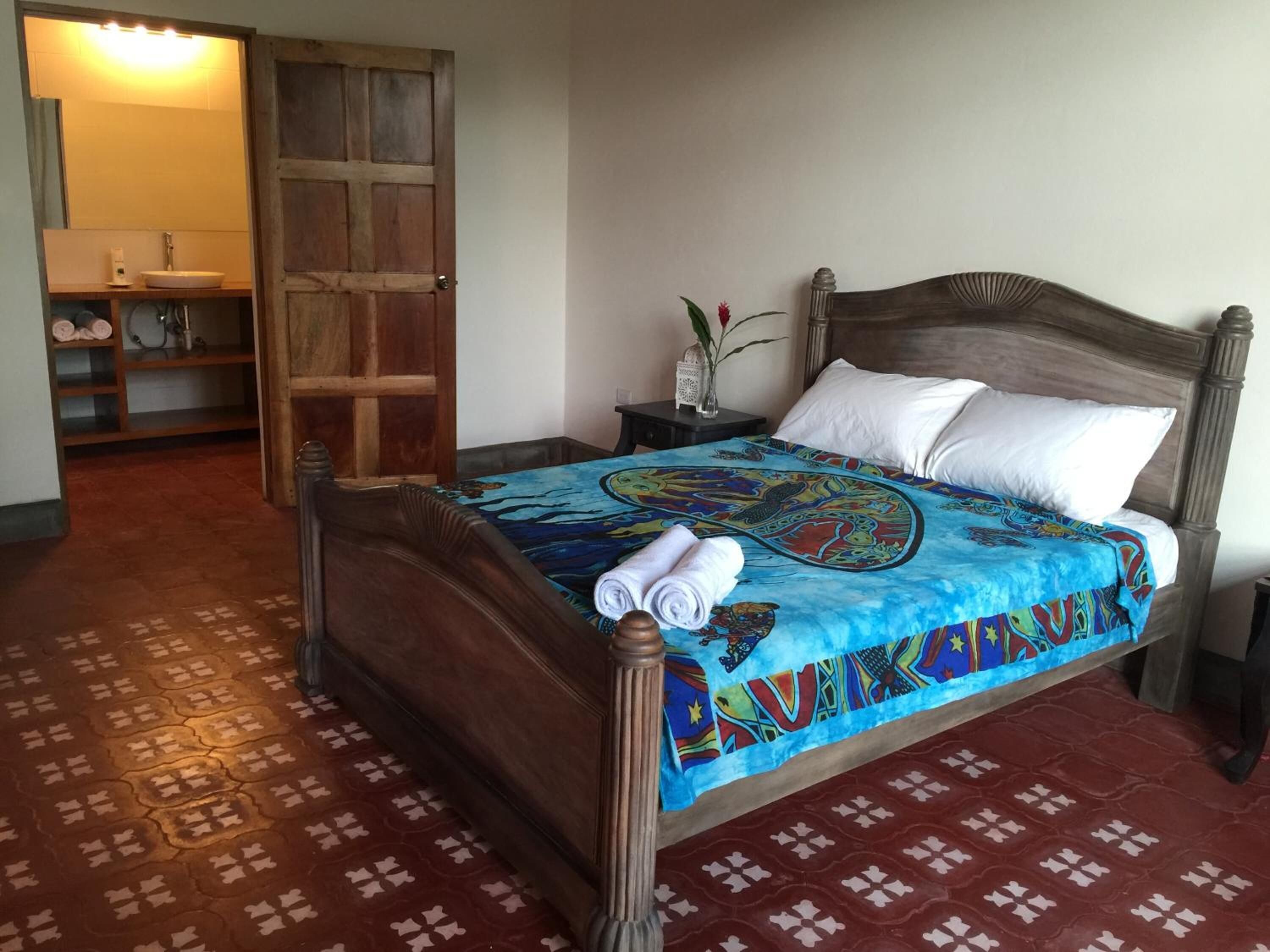 Une chambre avec un lit en bois, une table de chevet, un miroir et une salle de bain visible à travers une porte ouverte.
