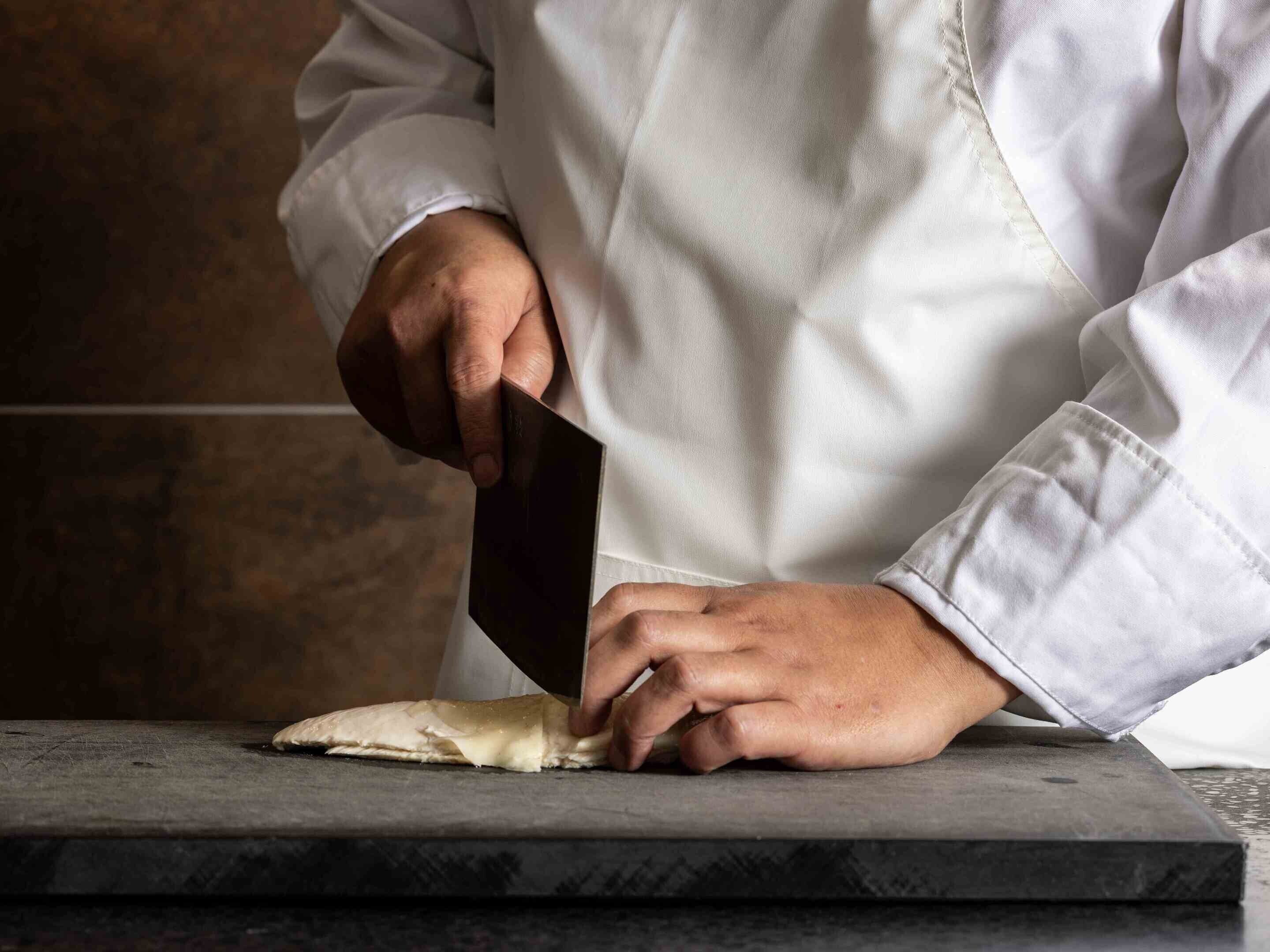 Una persona con delantal blanco de chef sosteniendo un cuchillo, preparando comida sobre una tabla de cortar.