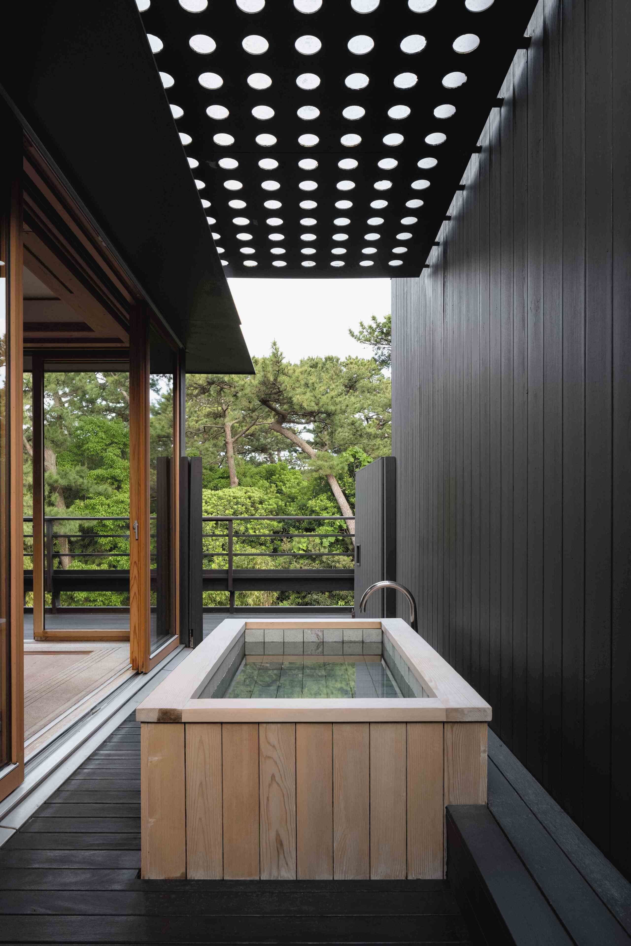 A modern outdoor hot tub area with a wooden deck, a pergola with circular cutouts, and a view of a lush garden.