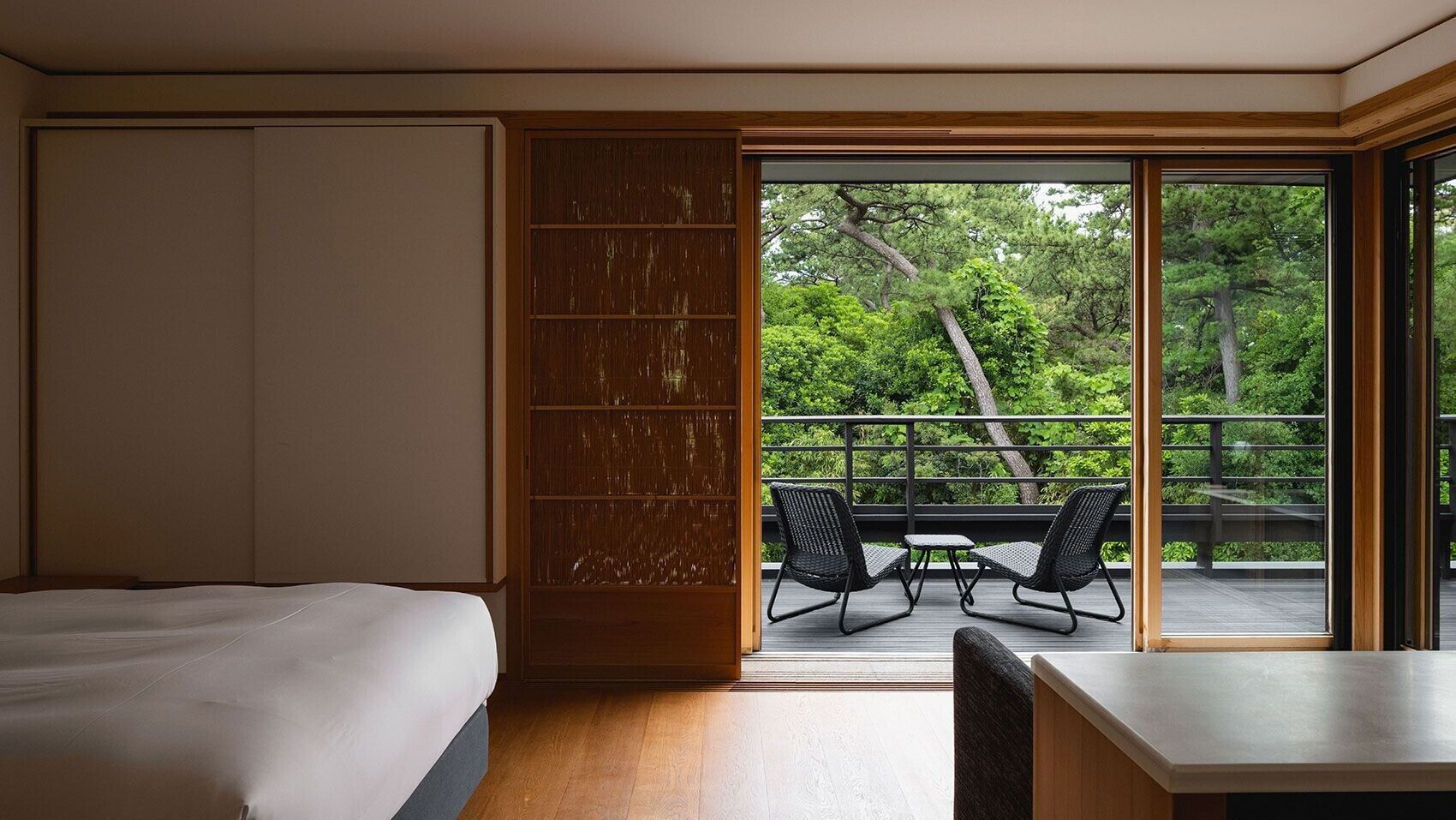 Habitación de hotel con cama, puerta corrediza a un balcón y vista a árboles.