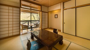 A traditional Japanese room with tatami flooring, a low table with chairs, a sliding door, and a large window with a view of trees. - Gokurakuji Onsen Arcadia Village (Hatsukaichi)