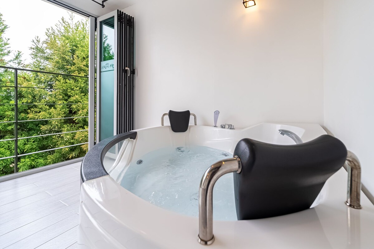 A modern bathroom with a bathtub, a shower, and a toilet. There is a glass door leading to a balcony with a view of trees.