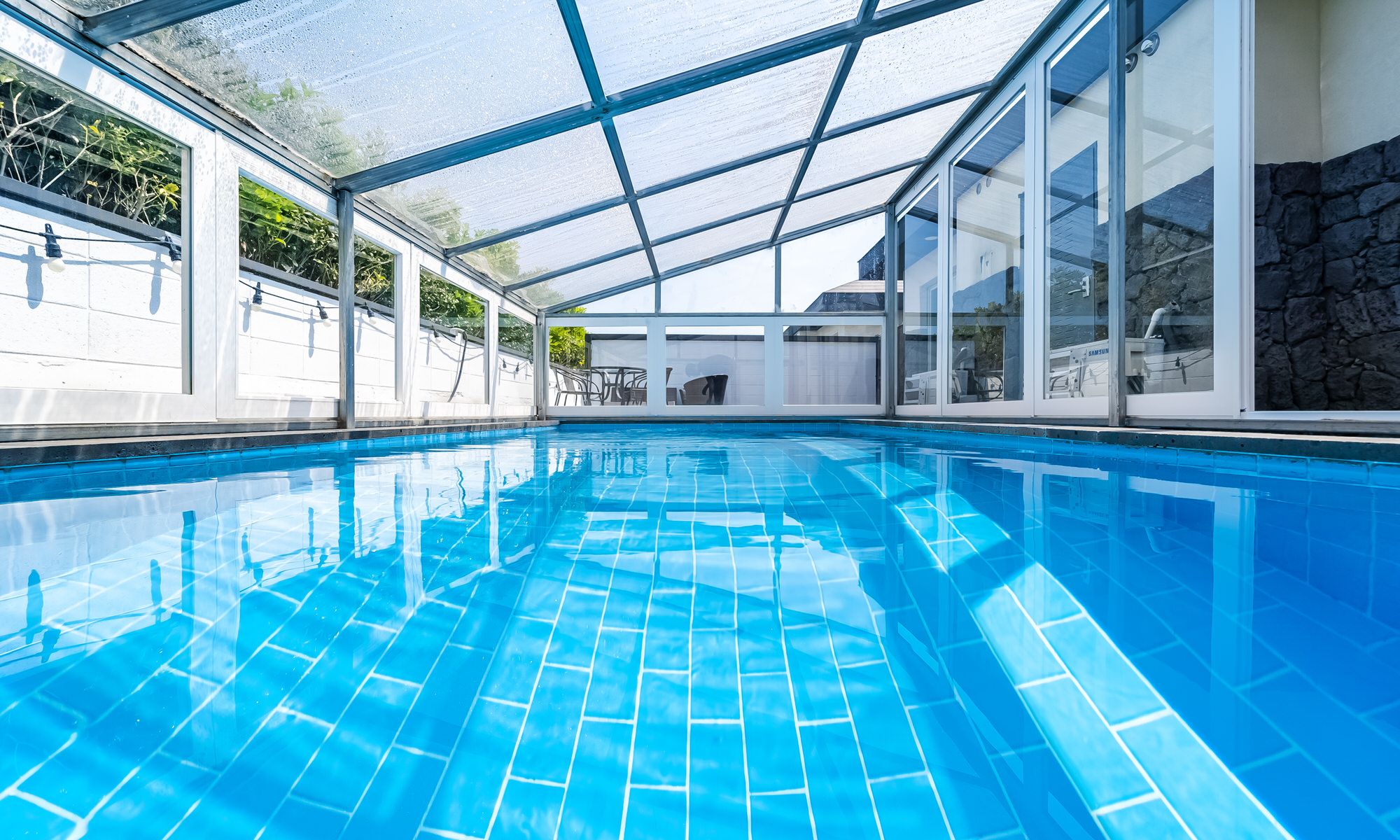 An indoor swimming pool with clear blue water, surrounded by glass walls and a metal frame structure.