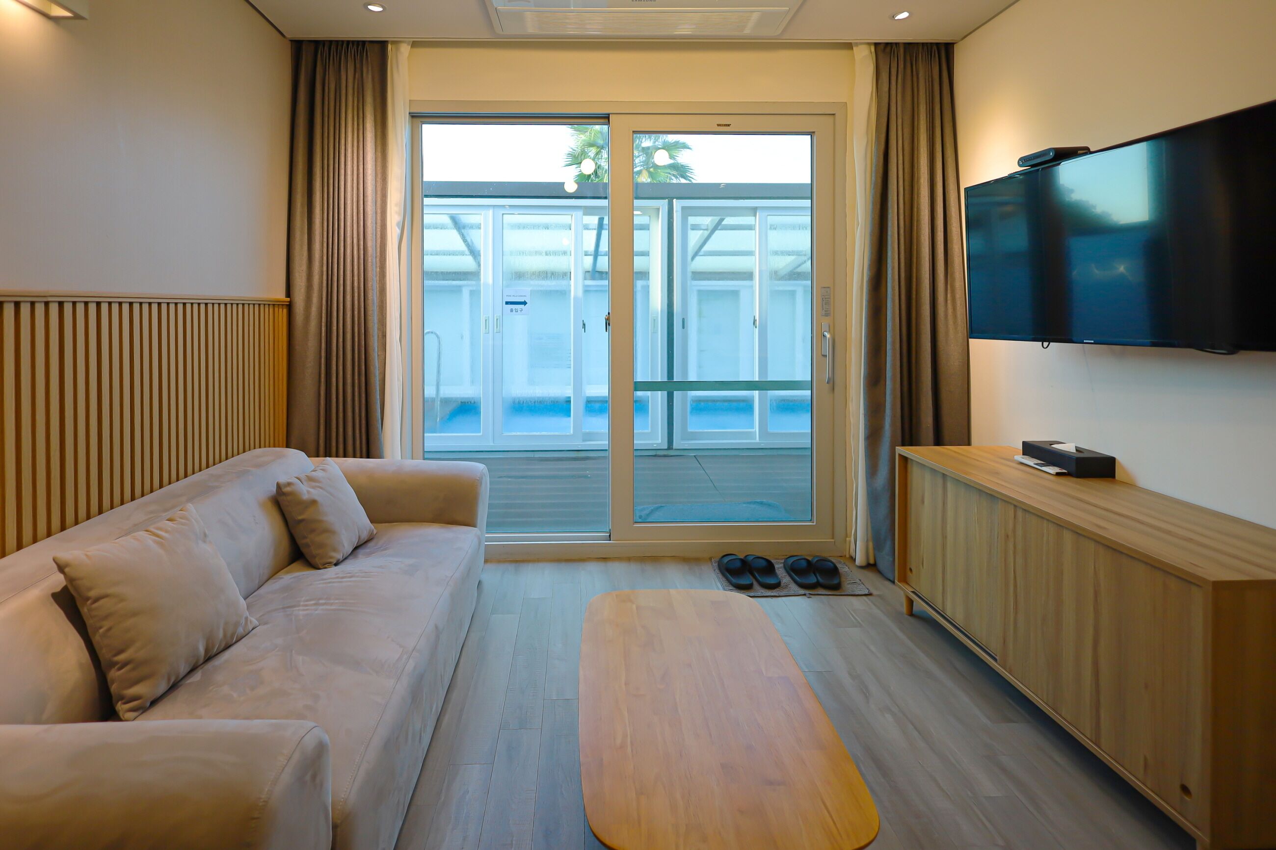 Habitación de hotel moderna con sofá, una mesa de centro de madera, televisión y puerta corredera de vidrio que da a una zona de piscina.