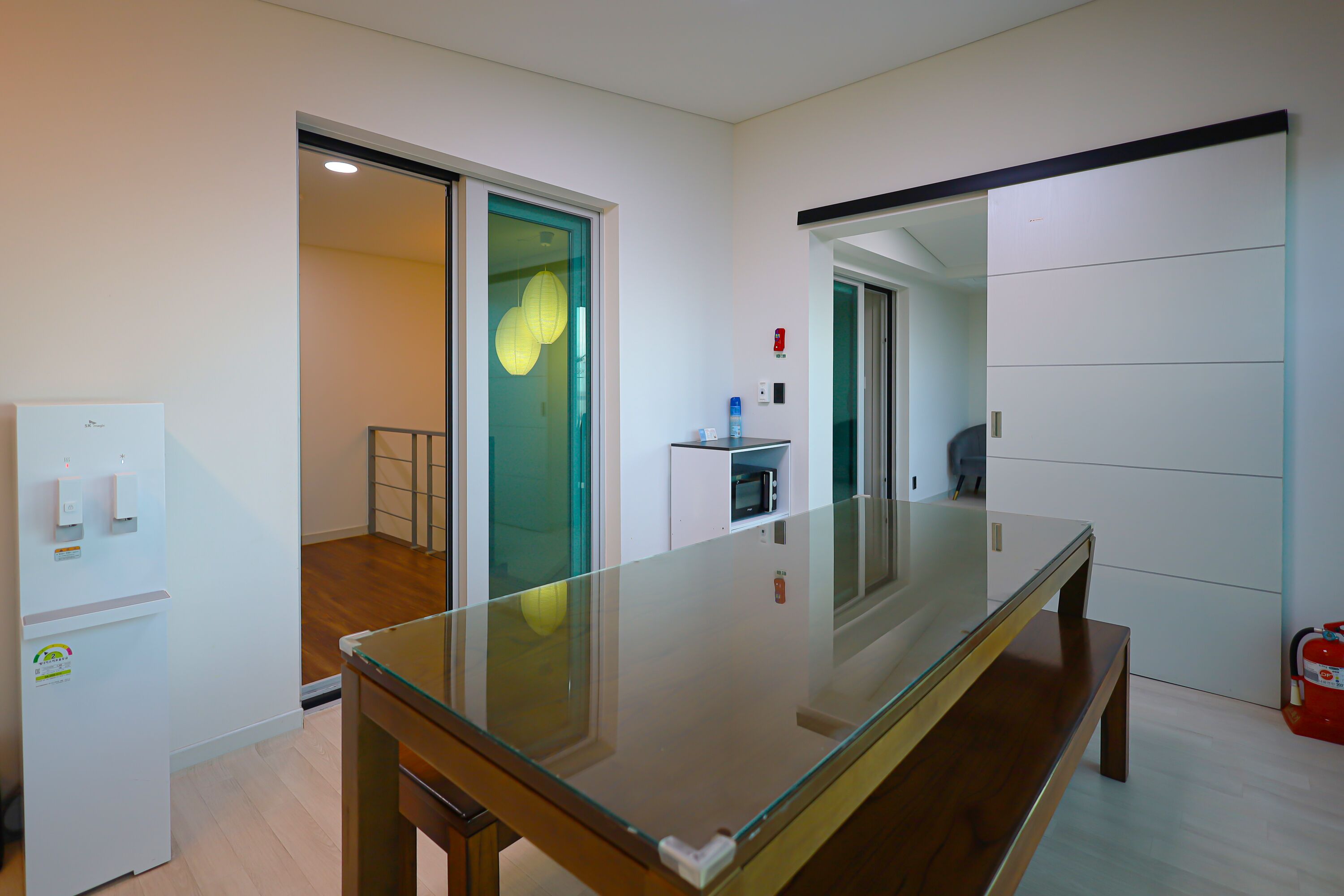 A modern hotel room with a glass-top table, a small refrigerator, a microwave, and a fire extinguisher.