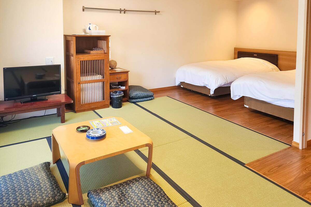 Um quarto com uma cama, televisão, armário de madeira, uma pequena mesa e piso de esteira tatame.