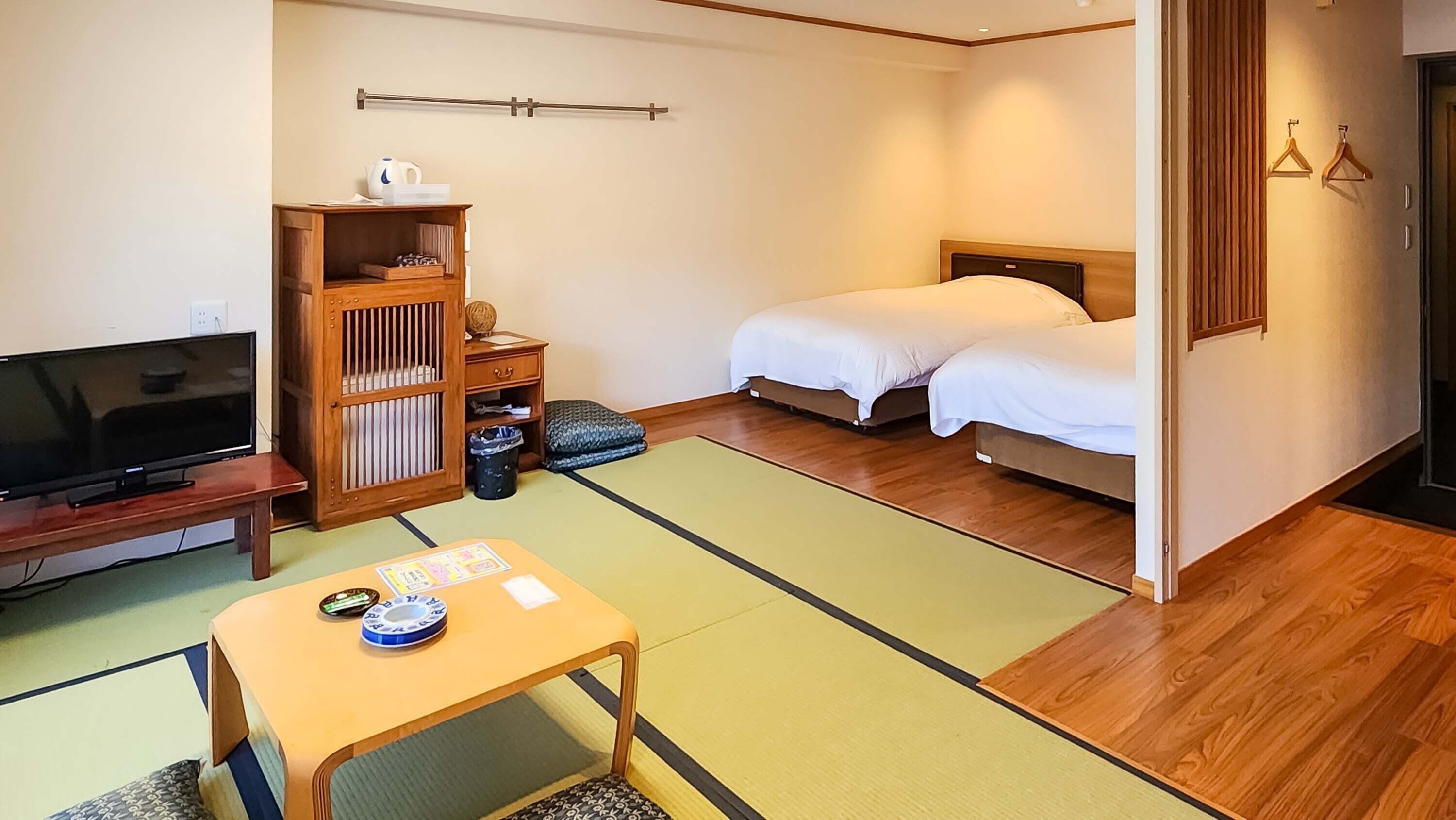 A hotel room with a bed, a television, a wooden cabinet, a tatami mat, and a small table with a cup and a book.