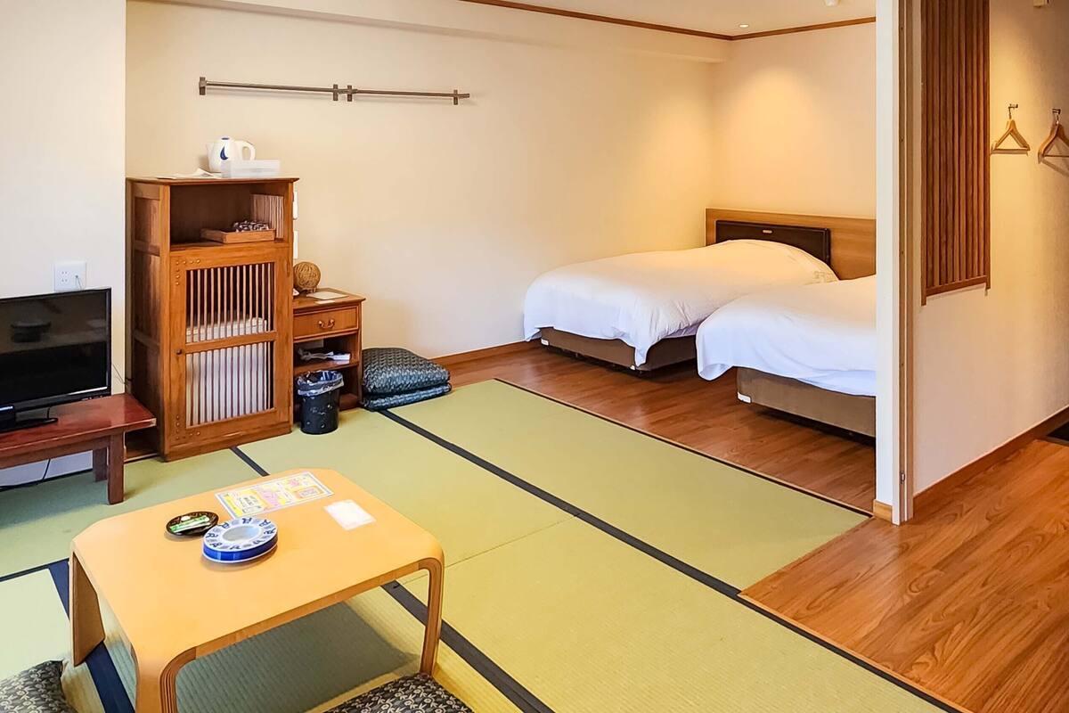 Quarto de hotel com cama, televisão, armário de madeira, esteira de tatame e uma pequena mesa com uma xícara e um livro.