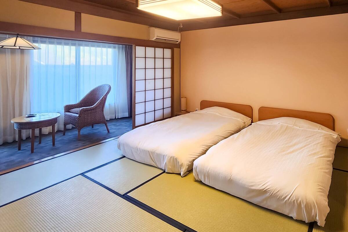 Um quarto tradicional em estilo japonês, com tatame, futon, divisória shoji e uma pequena mesa com cadeira.