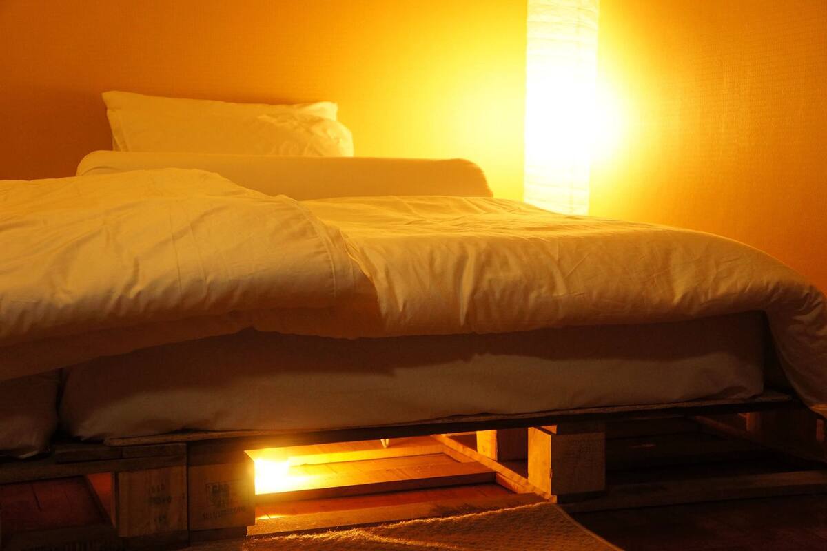 A bed with a wooden frame and white bedding, positioned under a window with a warm light source.