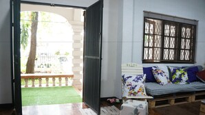 A room with a wooden floor, a sofa with floral cushions, a window with blinds, and a view of a garden outside. - Sleepy Oho (Chiang Mai)