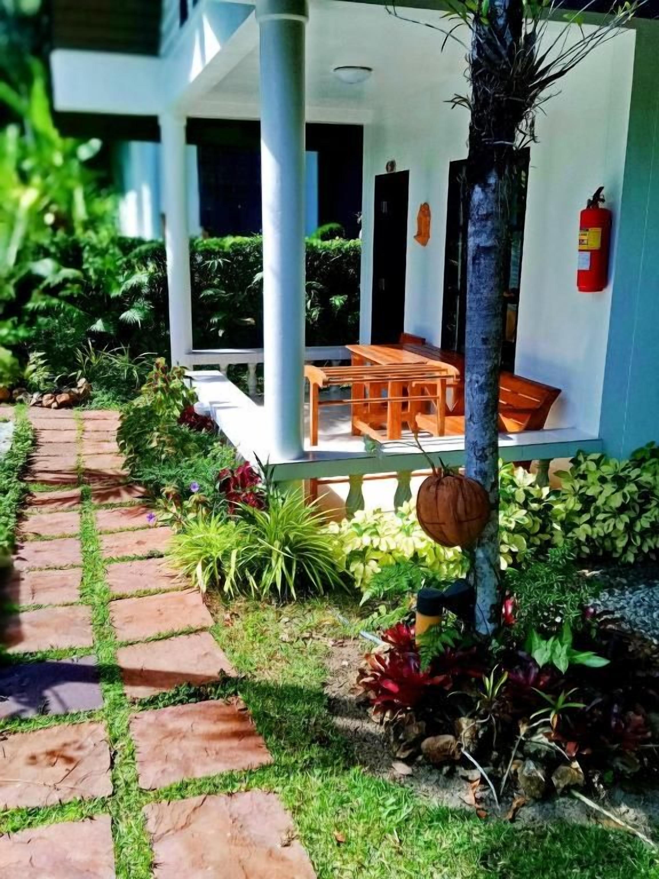 A covered outdoor seating area with a table and chairs, surrounded by lush greenery and a stone pathway.