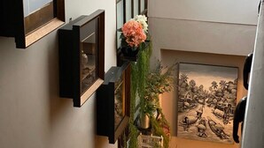 Ein Flur mit einer Holztreppe, einem an der Wand befestigten Regal mit gerahmten Bildern, einem Wandbehang mit floralem Muster und einem schwarz-weißen Gemälde in einem Rahmen.