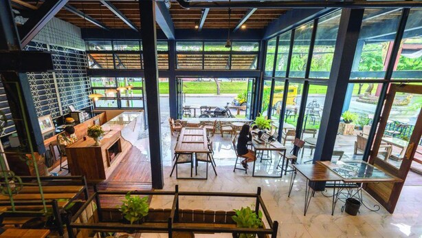 Un café amplio con grandes ventanales, mesas de madera y diferentes opciones de asientos.