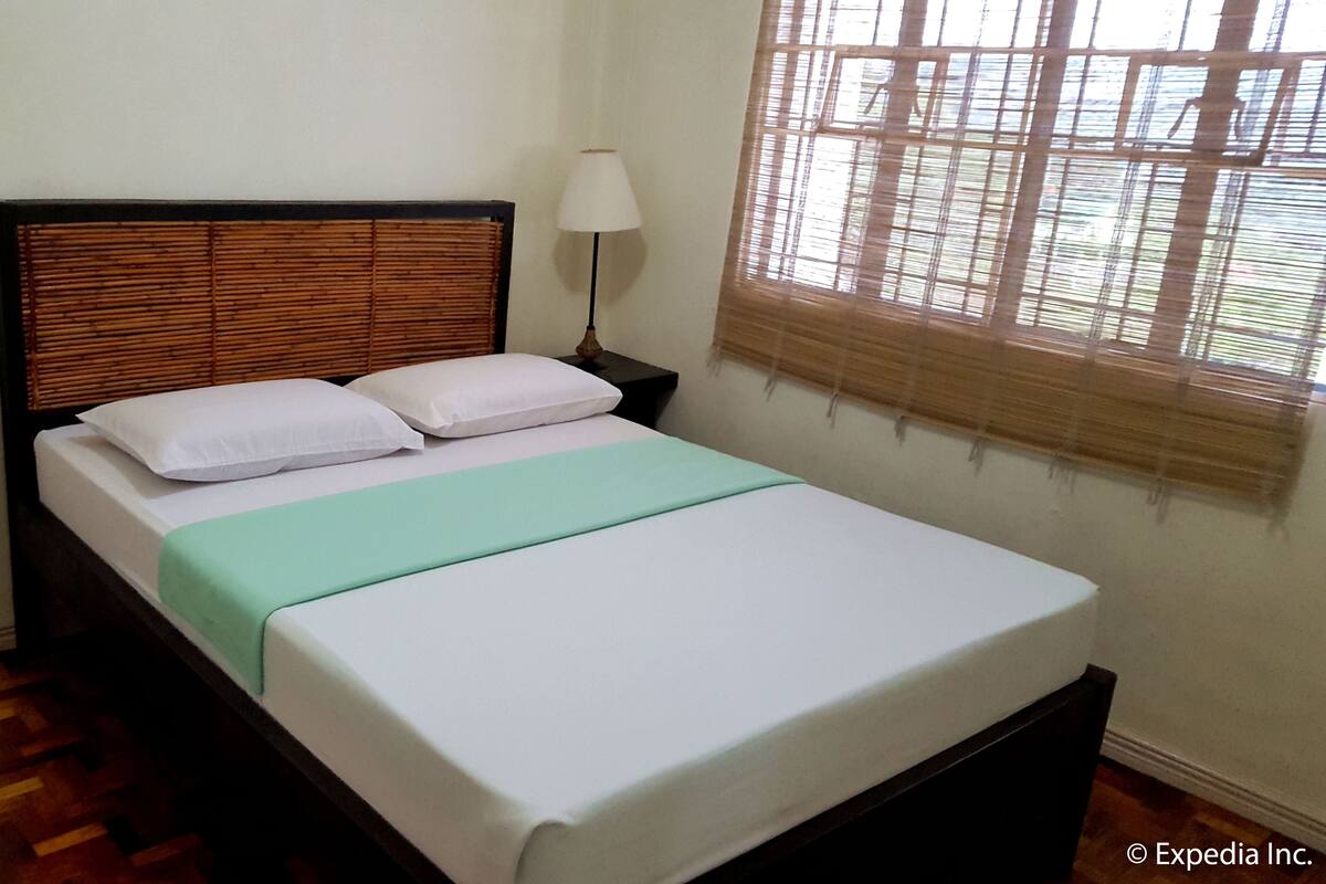 Una cama individual con cabecera de madera, dos almohadas blancas y una colcha verde en una habitación con suelo de madera y una ventana con persianas.