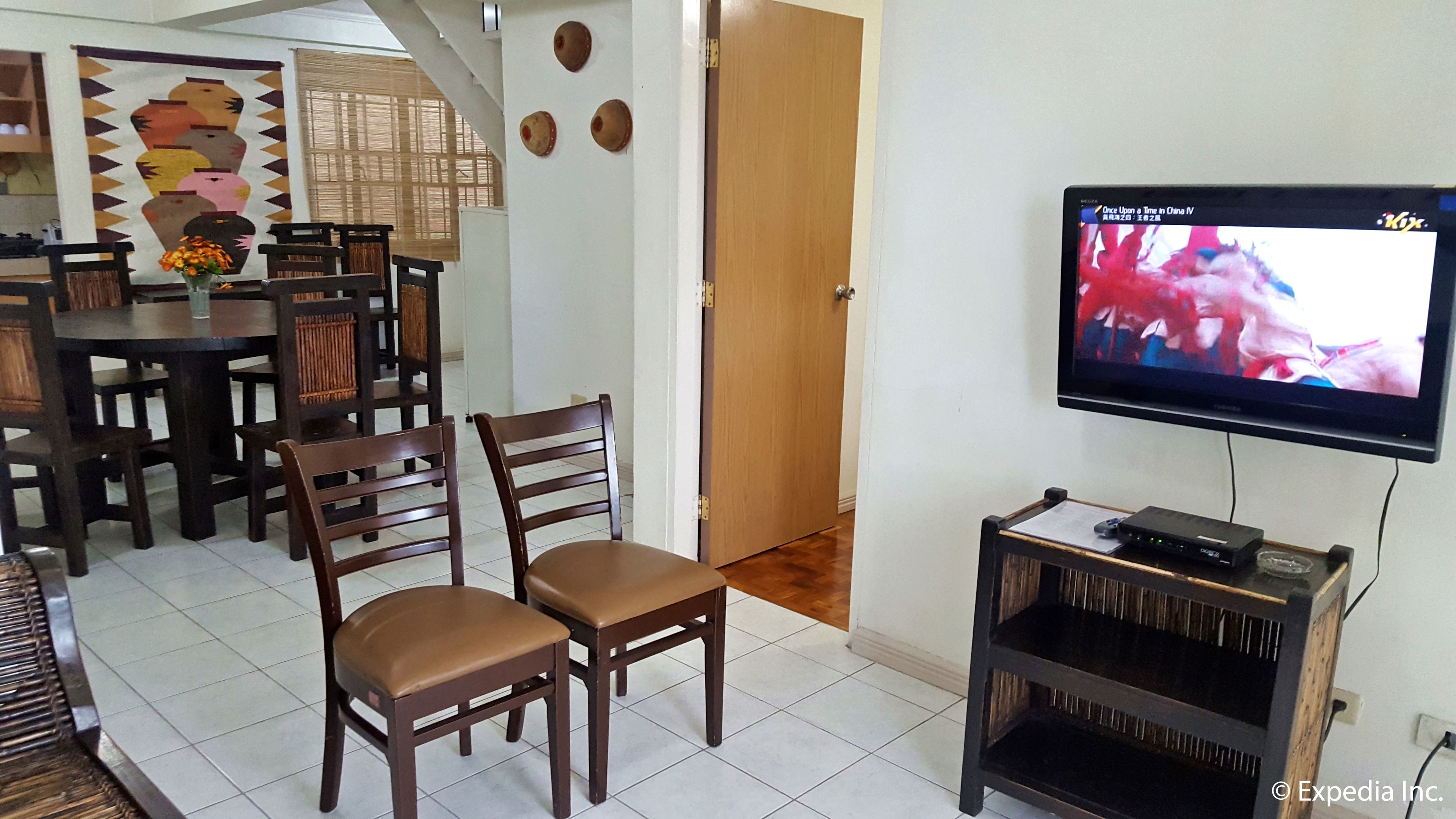 A living room with a TV mounted on the wall, wooden chairs, a dining table, and a decorative wall hanging.