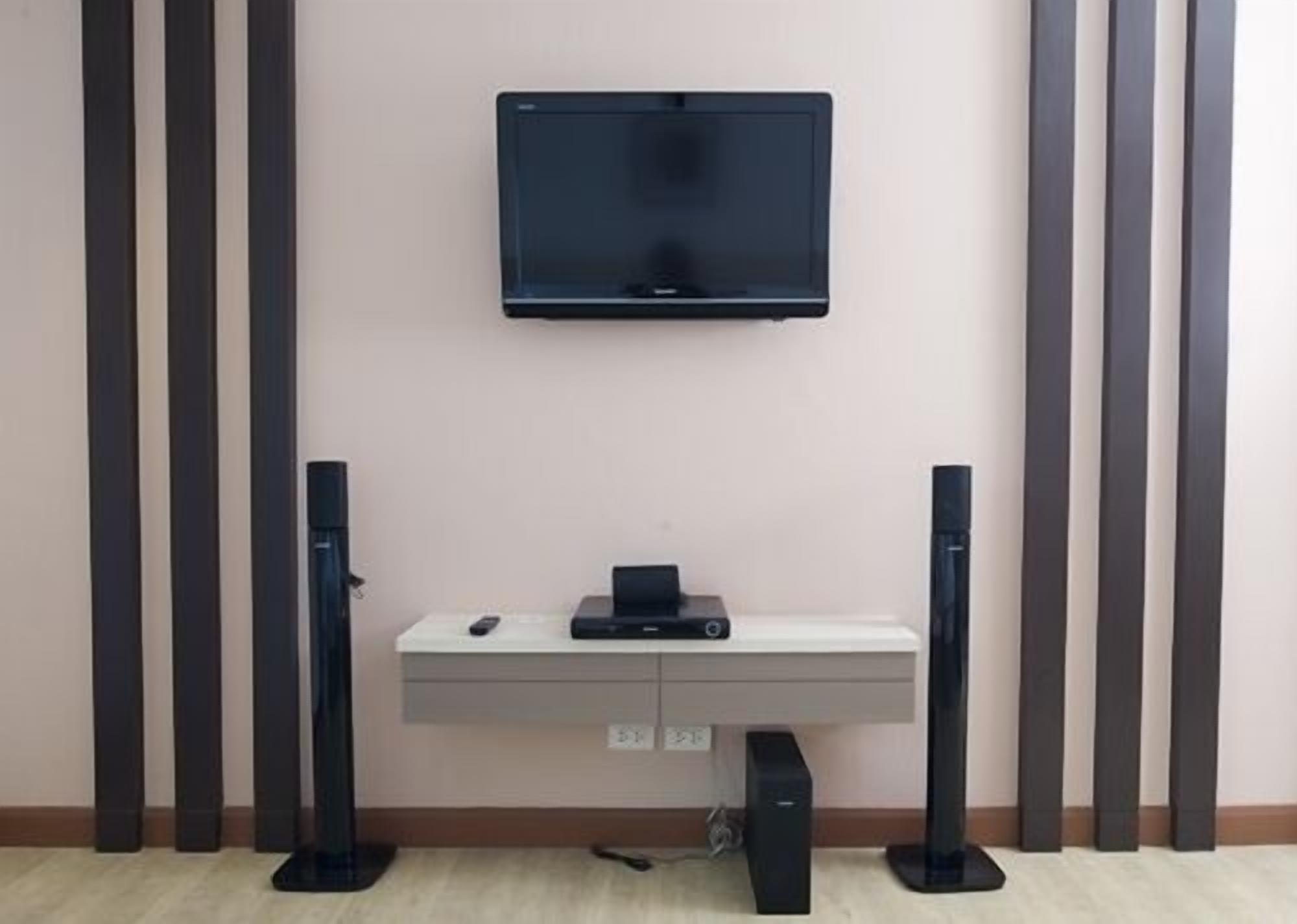 A room with a mounted television on the wall, a white console table with a remote and a small electronic device, and two black speakers on either side.