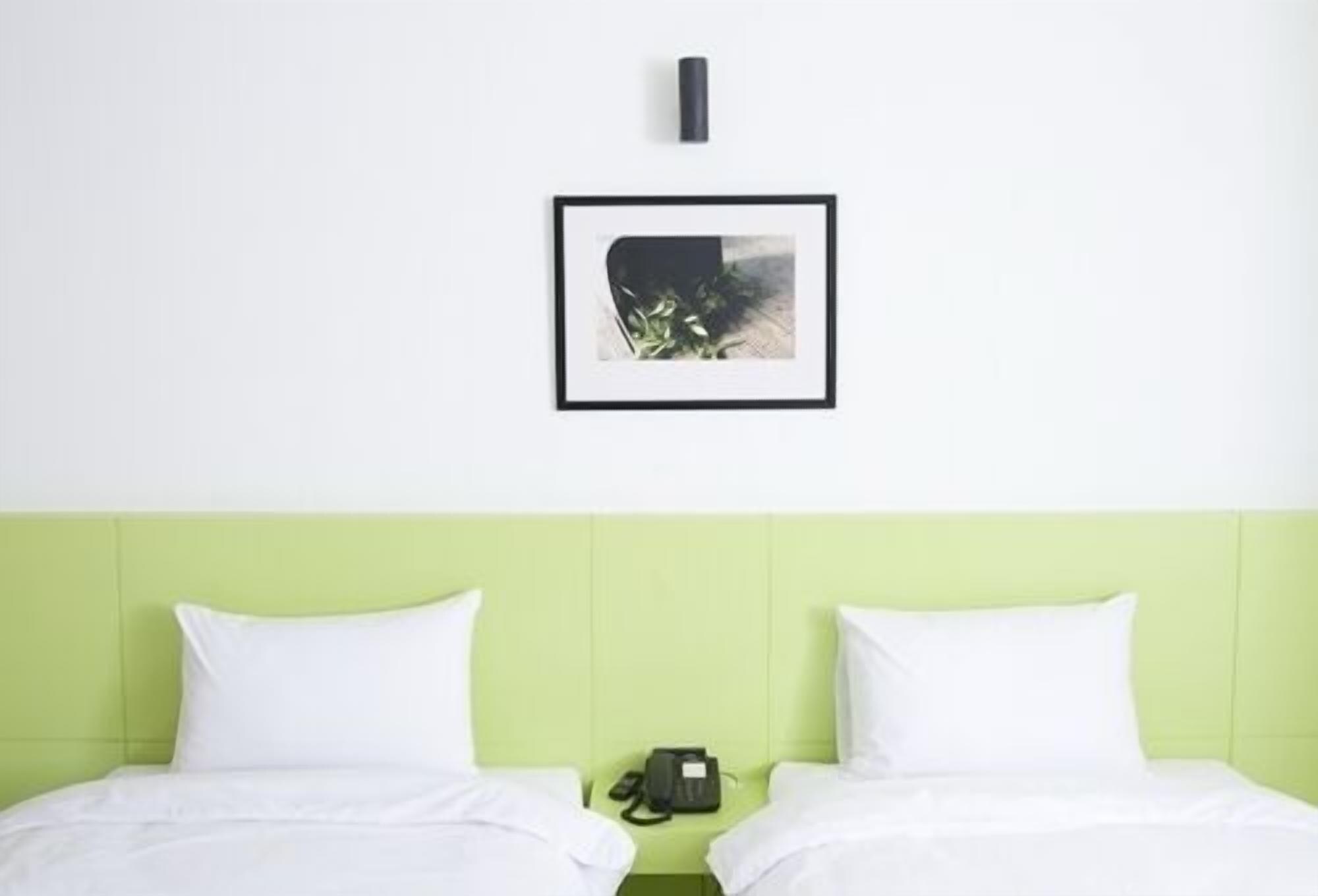 Habitación de hotel con cabecera verde, dos camas blancas, un cuadro en la pared y una mesita con teléfono y lámpara.