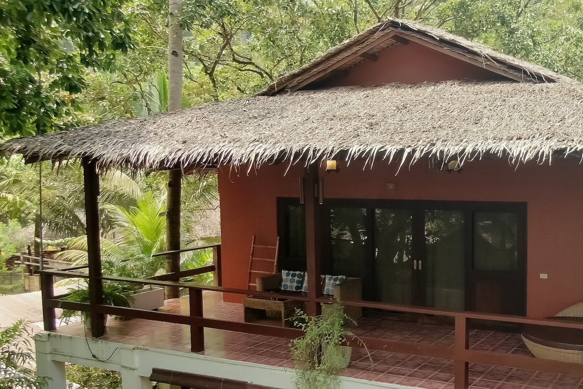 A small, single-story building with a thatched roof, a balcony with seating, and a view of lush greenery.