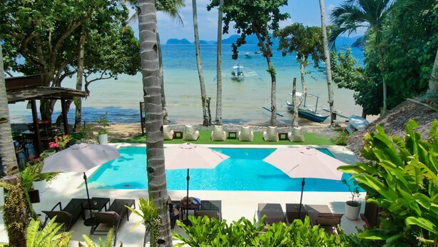 A pool area with lounge chairs and umbrellas, overlooking a beach with boats and palm trees. - Last Frontier Beach Resort - Adults Only (El Nido)