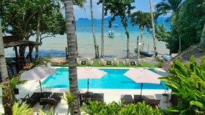 A pool area with lounge chairs and umbrellas, overlooking a beach with boats and palm trees.