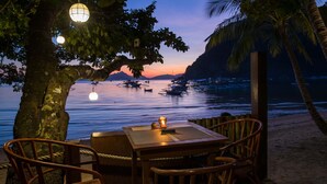 A table and chairs under a tree by a beach with a view of boats and a sunset. - Last Frontier Beach Resort - Adults Only (El Nido)