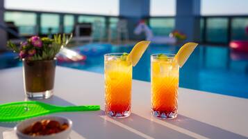 Dos vasos de una bebida en capas con rodajas de limón, un bol de frutos secos y una bandeja verde sobre una mesa junto a una piscina.