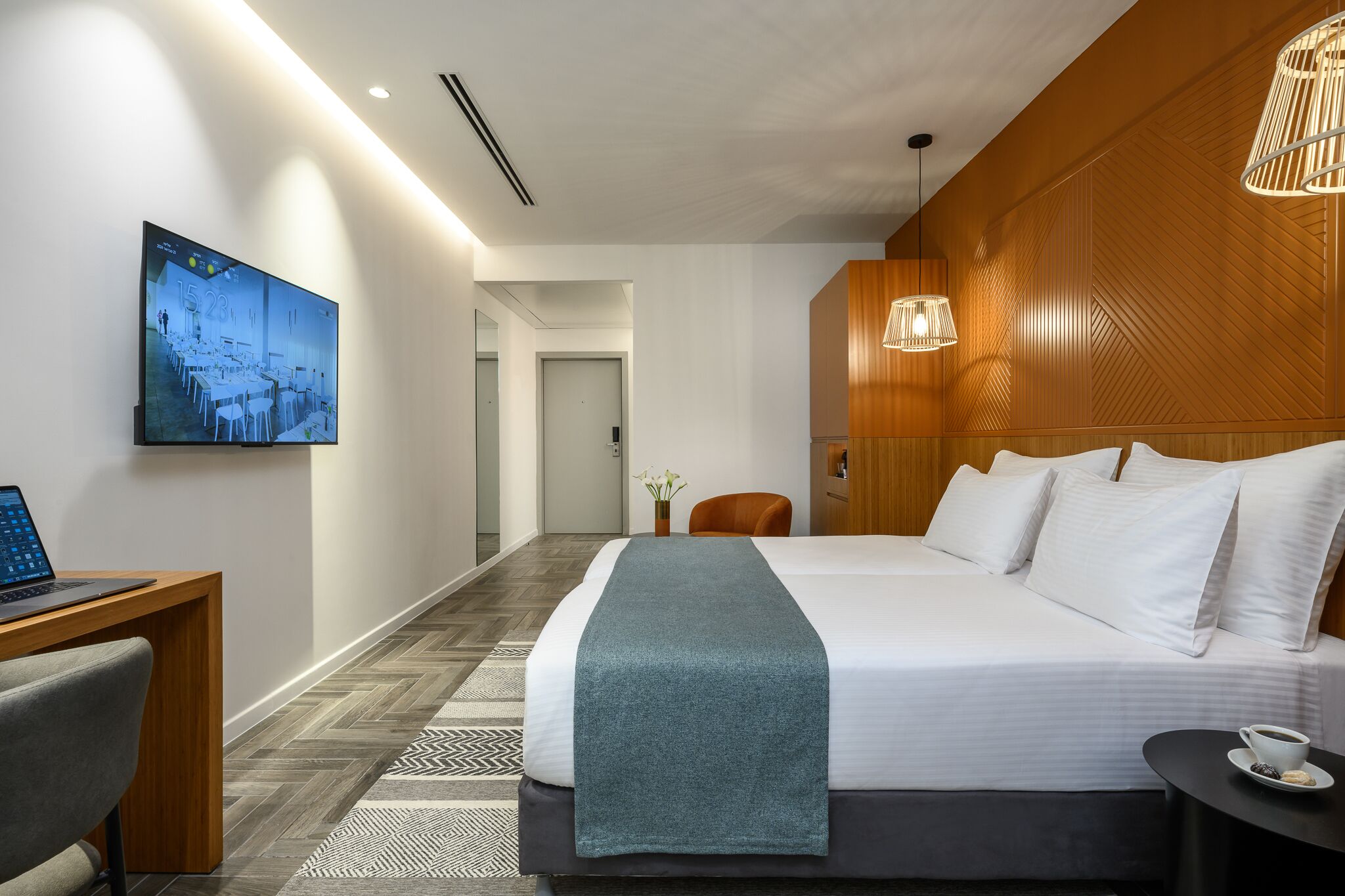 Et moderne hotelværelse med en stor seng, et skrivebord med en bærbar computer, et fjernsyn på væggen og en træpanelfront.