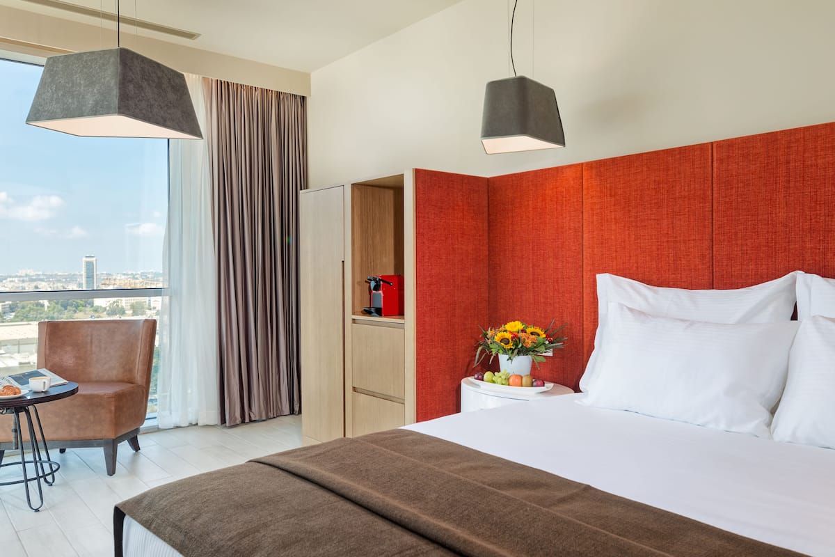 Une chambre d’hôtel avec un lit, une chaise, un bureau, un mur d’accent rouge et une vue sur la ville.