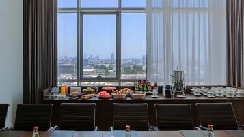 A conference room with a city view, equipped with a buffet table, chairs, and a coffee station.