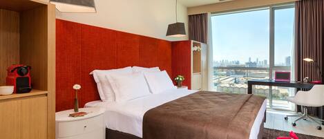 Habitación de hotel con una cama grande, un escritorio con silla, vista a la ciudad y una pared de acento roja.