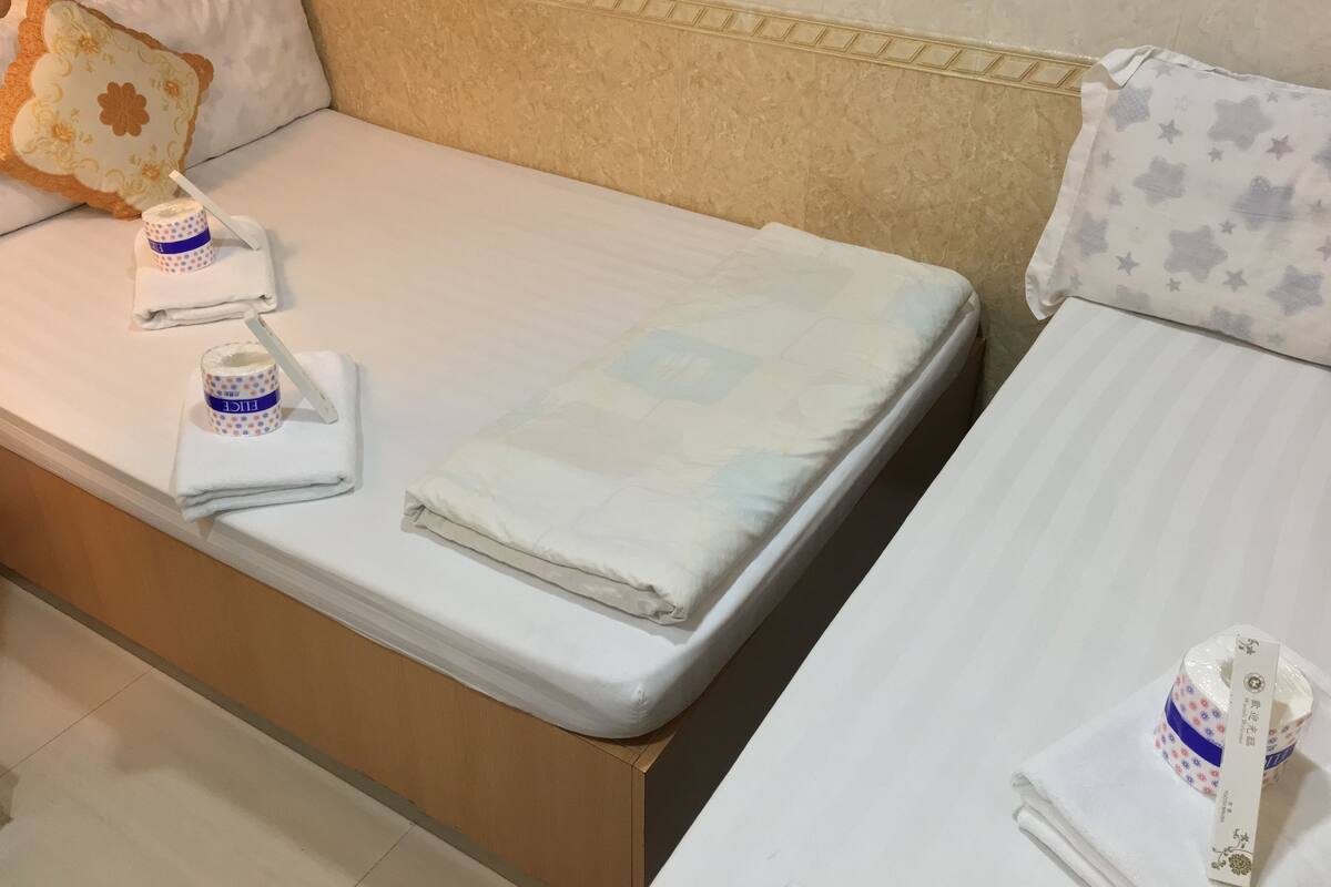 A hotel room with two beds, each with neatly folded white linens and a pillow. There are two decorative pillows with floral patterns and a star pattern. A small table between the beds holds a roll of toilet paper and a bottle of lotion.