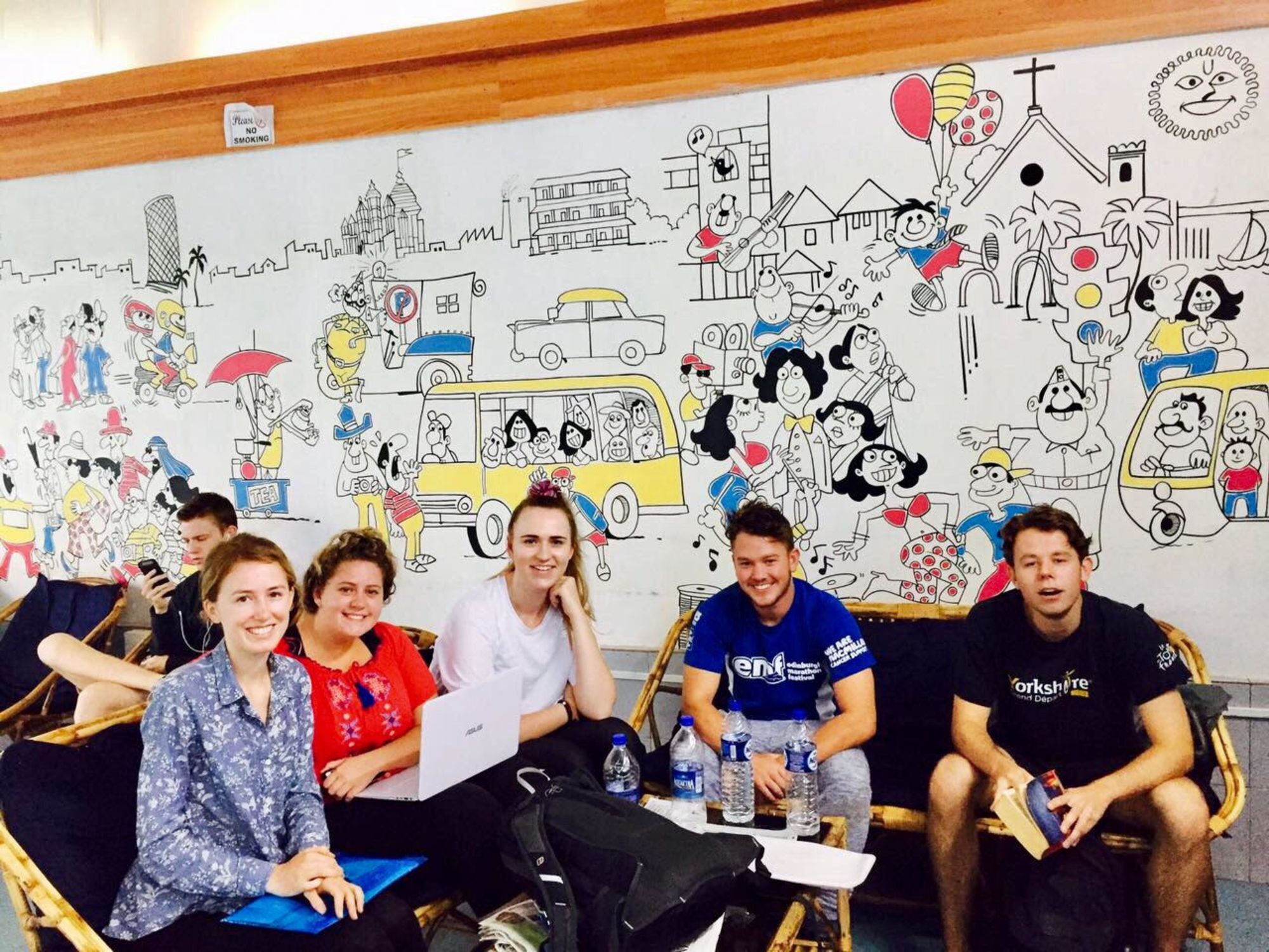 Un grup de persones assegudes en una sala amb un mural gran i colorit al mur que representa diverses escenes i personatges.
