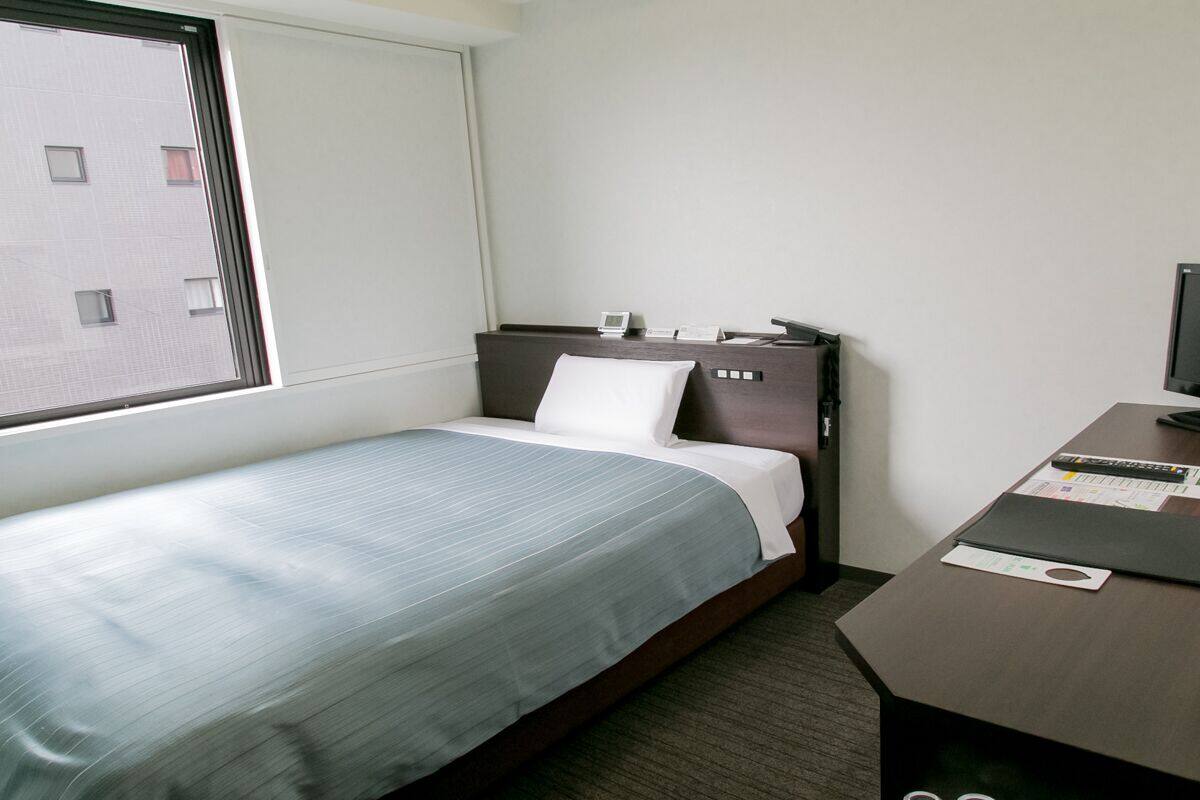Hotelkamer met een enkel bed, een bureau met een computer en een raam met uitzicht op gebouwen.