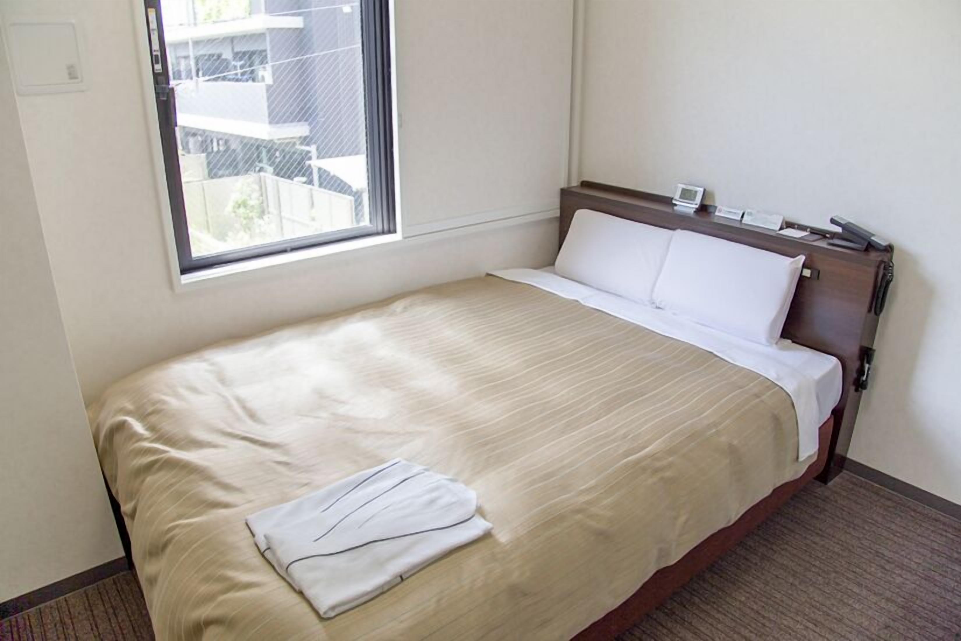 Une chambre d’hôtel avec un lit simple, une fenêtre avec des stores, une table de chevet avec une lampe et un thermostat fixé au mur.