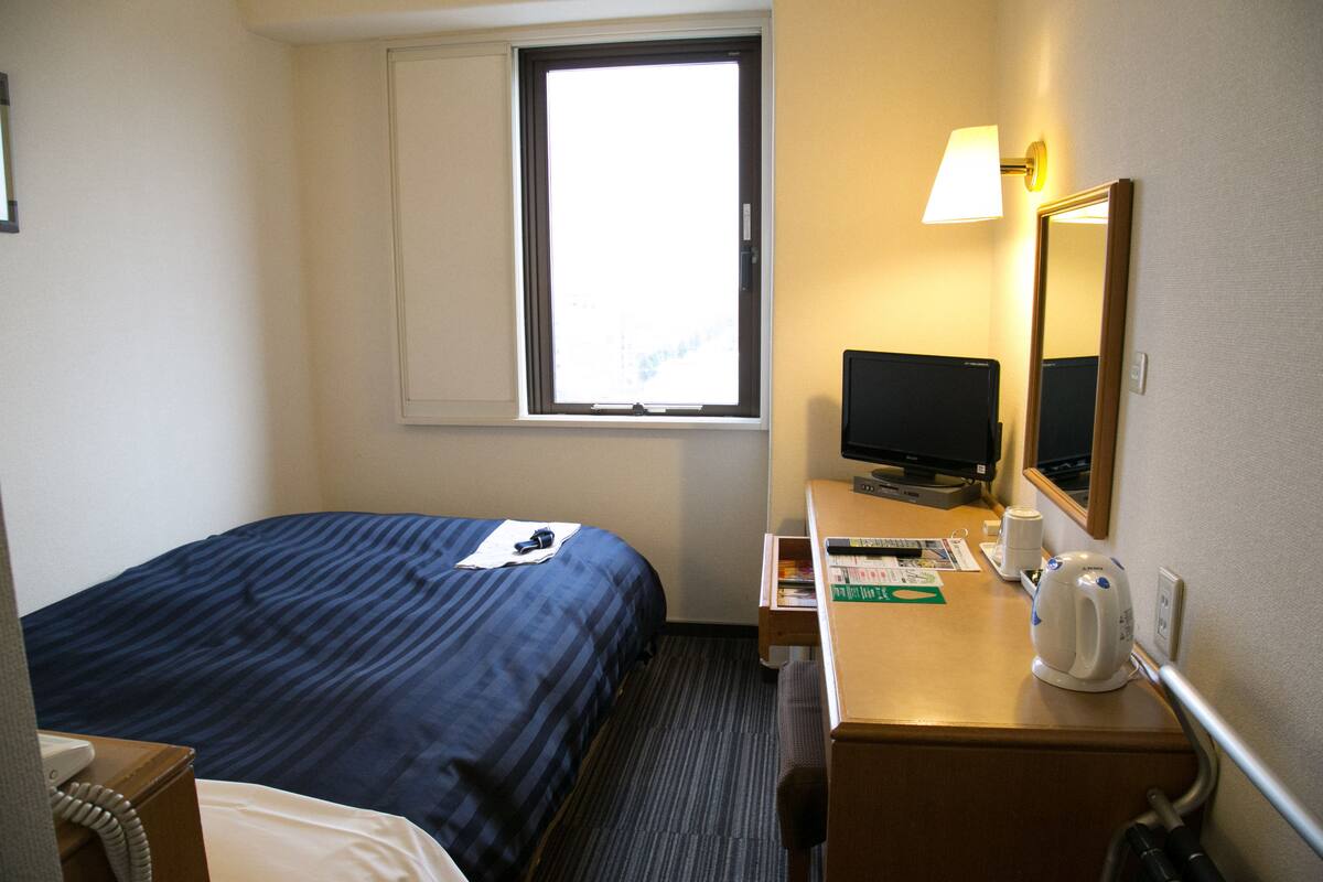 Een hotelkamer met een bed, een bureau met een computer, een waterkoker en een raam.